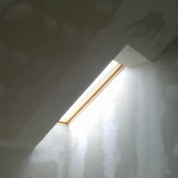 Wnętrze poddasza z oknem dachowym w trakcie prac wykończeniowych, ściany i sufit pokryte gładzią gipsową, widoczne łączenia płyt gipsowo-kartonowych.